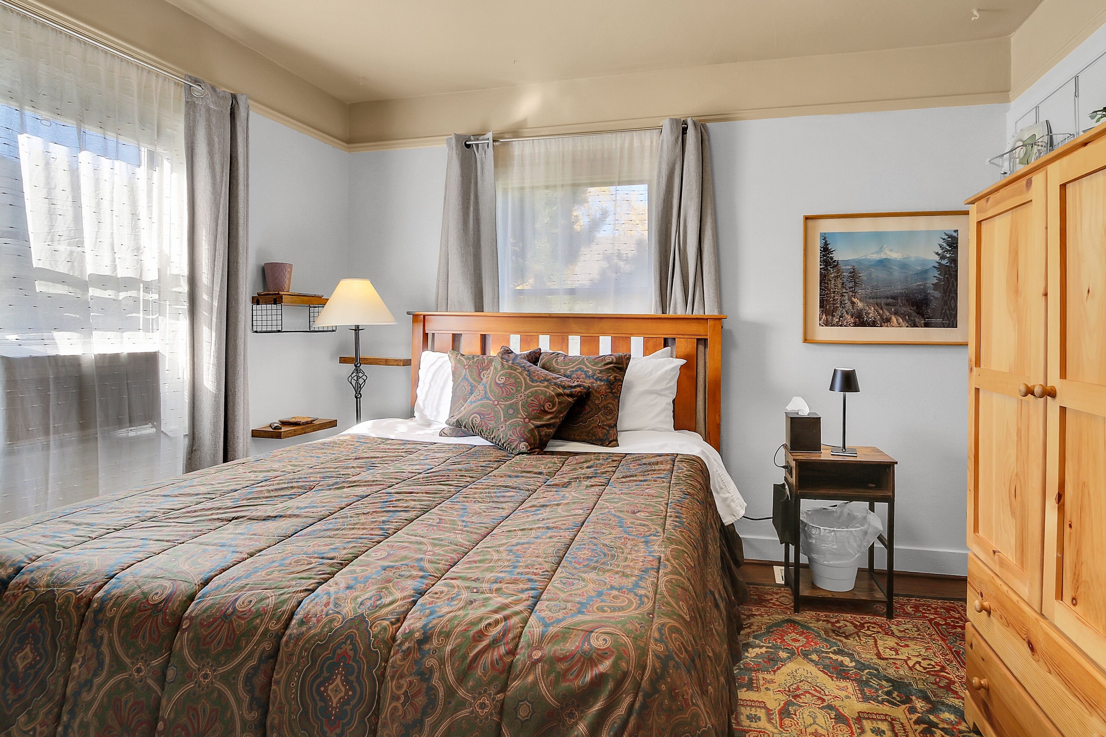 A neatly made bed with patterned bedding, a bedside table with a lamp, a small trash bin, and a wooden cabinet.
