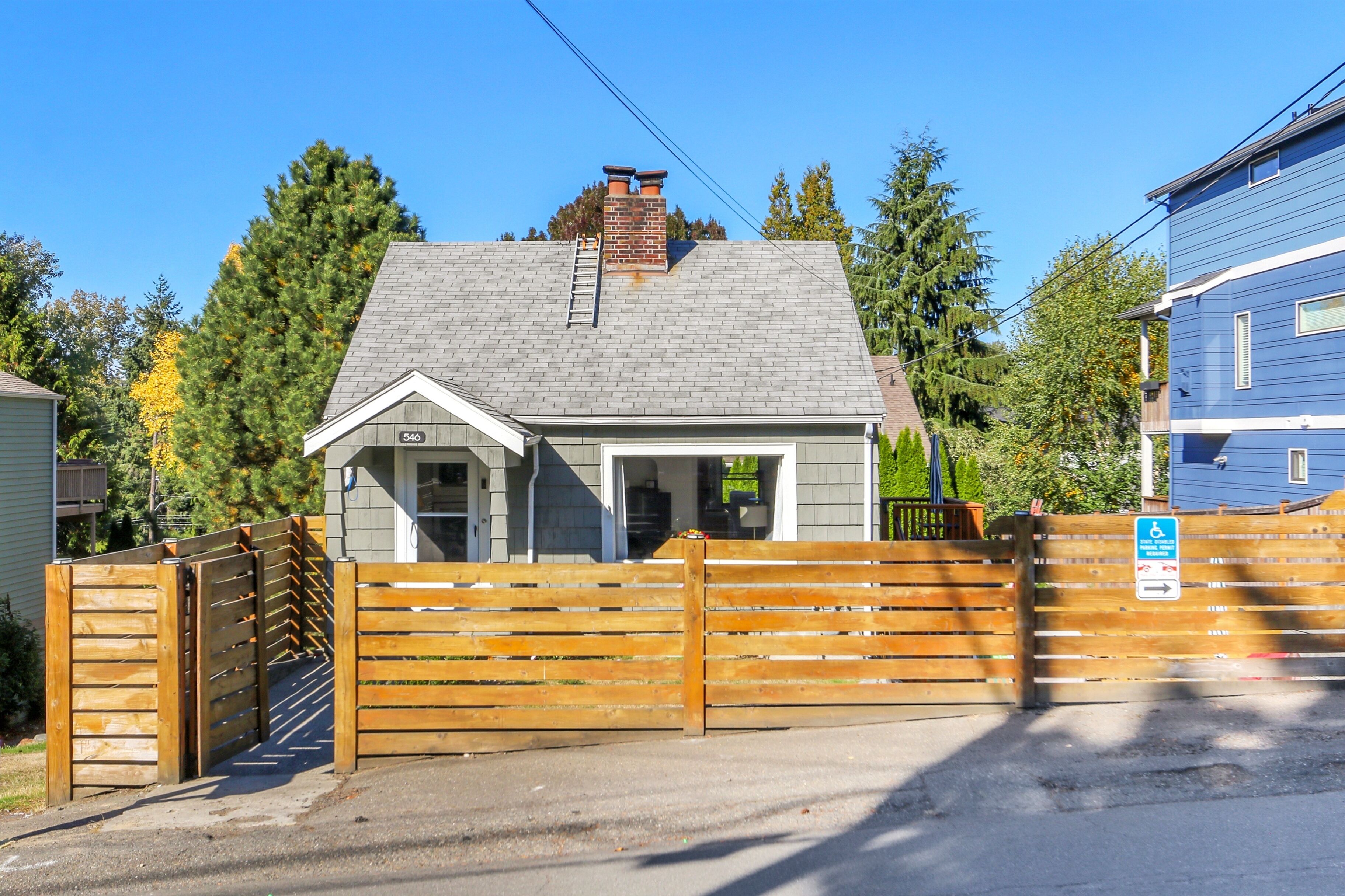 A small house with a grey roof, a wooden gate, and a blue sign indicating a wheelchair-accessible entrance.