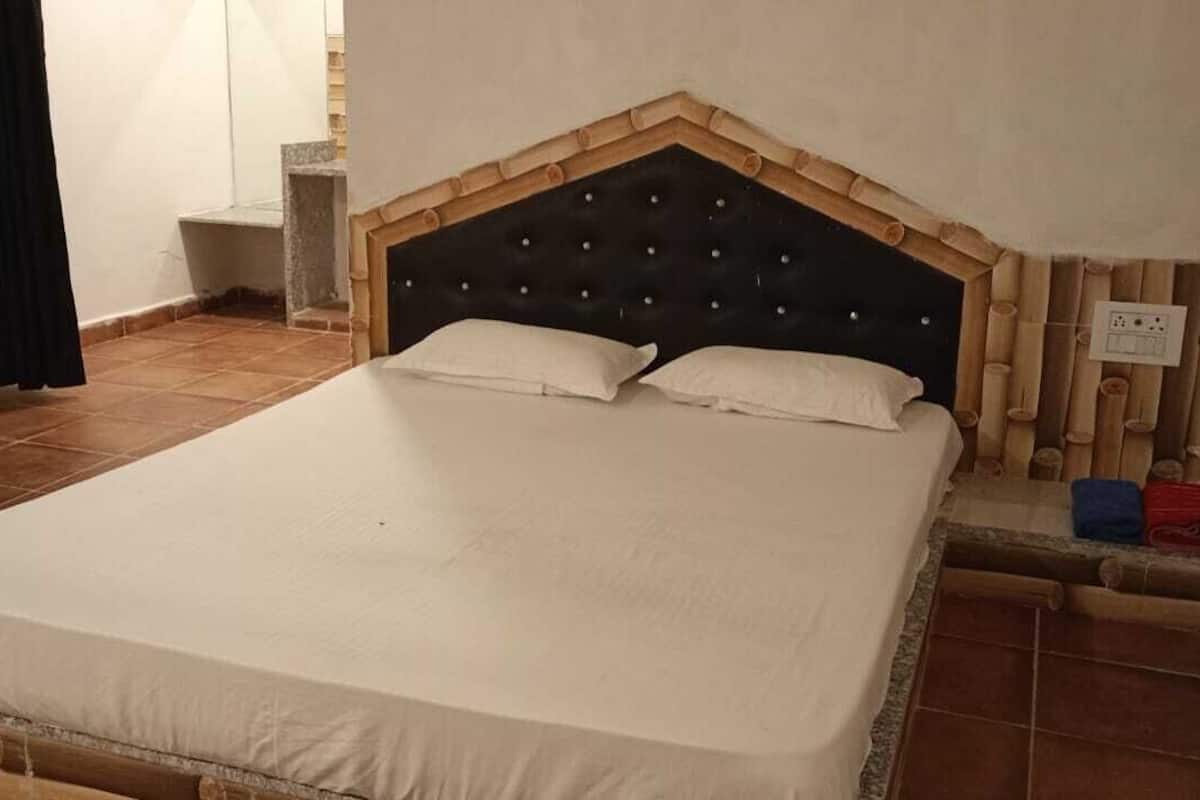 A single bed with a wooden headboard and two pillows, a small bedside table, and a power outlet on the wall.