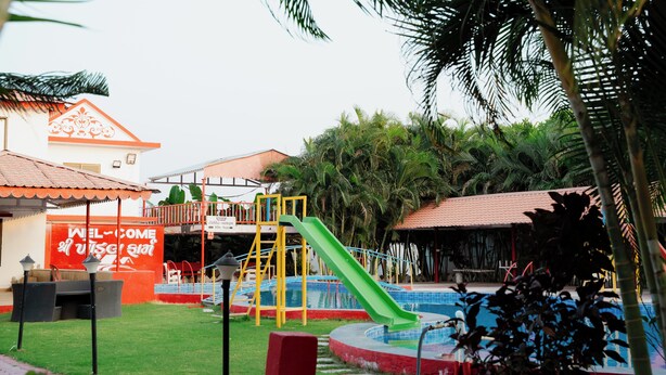 A playground with a slide and a pool, surrounded by palm trees and a building with a red sign. - khodal Resort (Girgadhada)