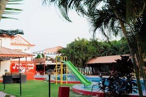 A playground with a slide and a pool, surrounded by palm trees and a building with a red sign. - khodal Resort (Girgadhada)