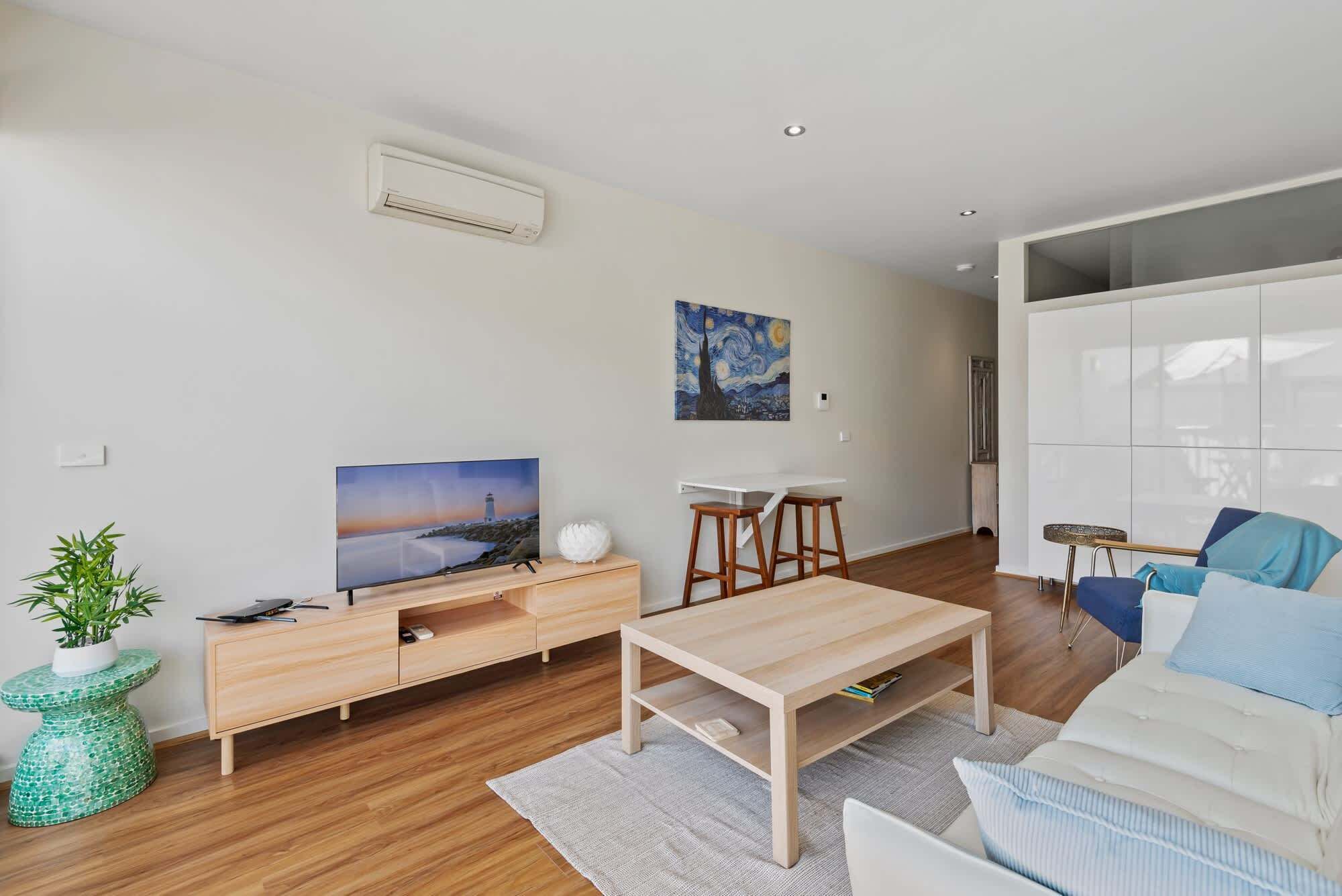 A modern living room with a flat-screen TV, a painting, a white sofa, a coffee table, and a dining area with stools.