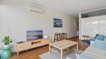 A modern living room with a flat-screen TV, a painting, a white sofa, a coffee table, and a dining area with stools.