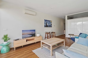 A modern living room with a flat-screen TV, a painting, a white sofa, a coffee table, and a dining area with stools.