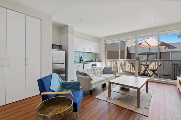 A modern living room with a sofa, a coffee table, and a dining table. There is a kitchen area with cabinets and a microwave. The room has large windows and a balcony with outdoor furniture.