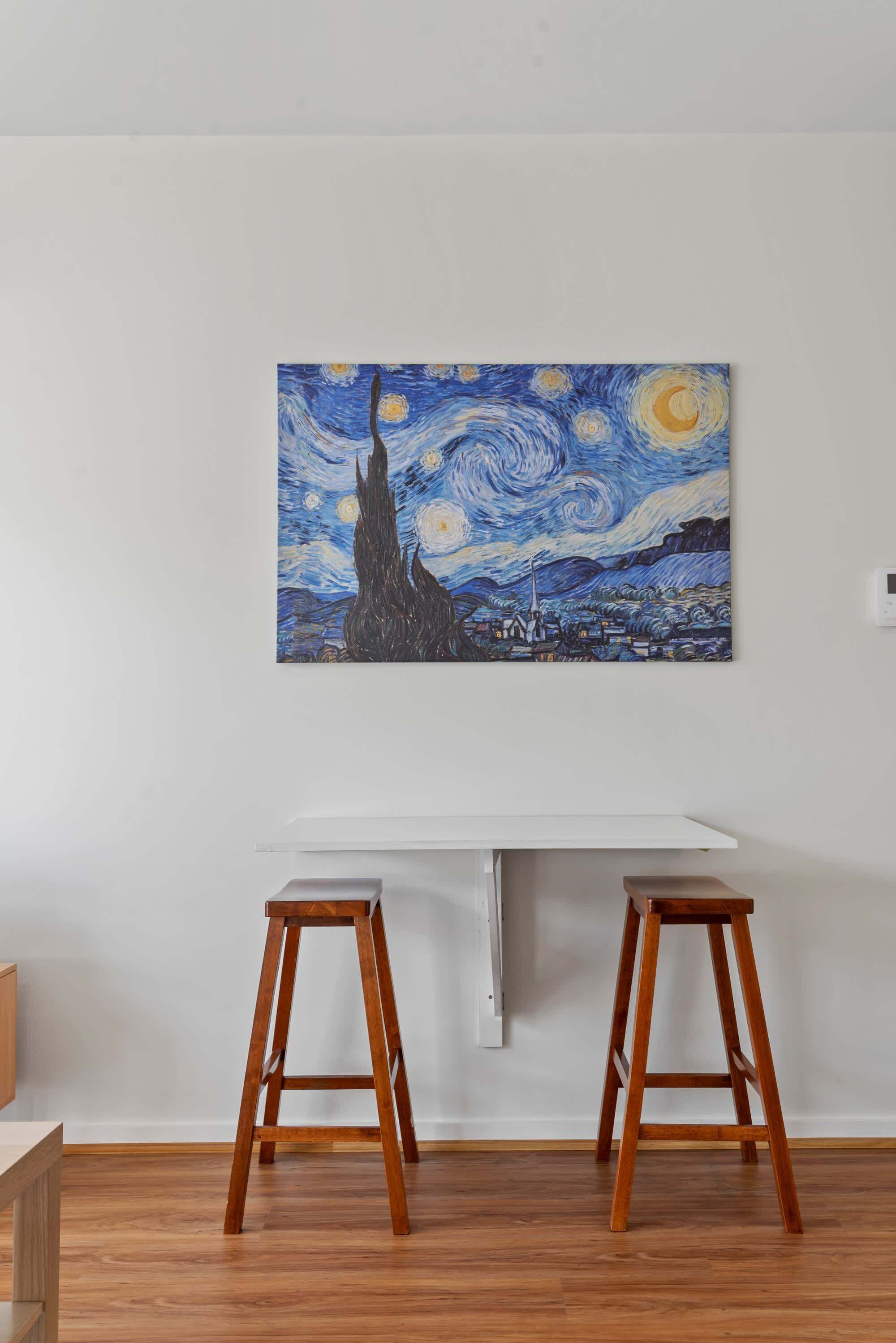 Une pièce avec un bureau blanc, deux tabourets en bois et un tableau accroché au mur.