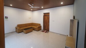 A room with a wooden ceiling, a ceiling fan, a brown sectional sofa, a small table, a wooden door, and a wooden cabinet. - HIMANI HERITAGE (Madikeri)