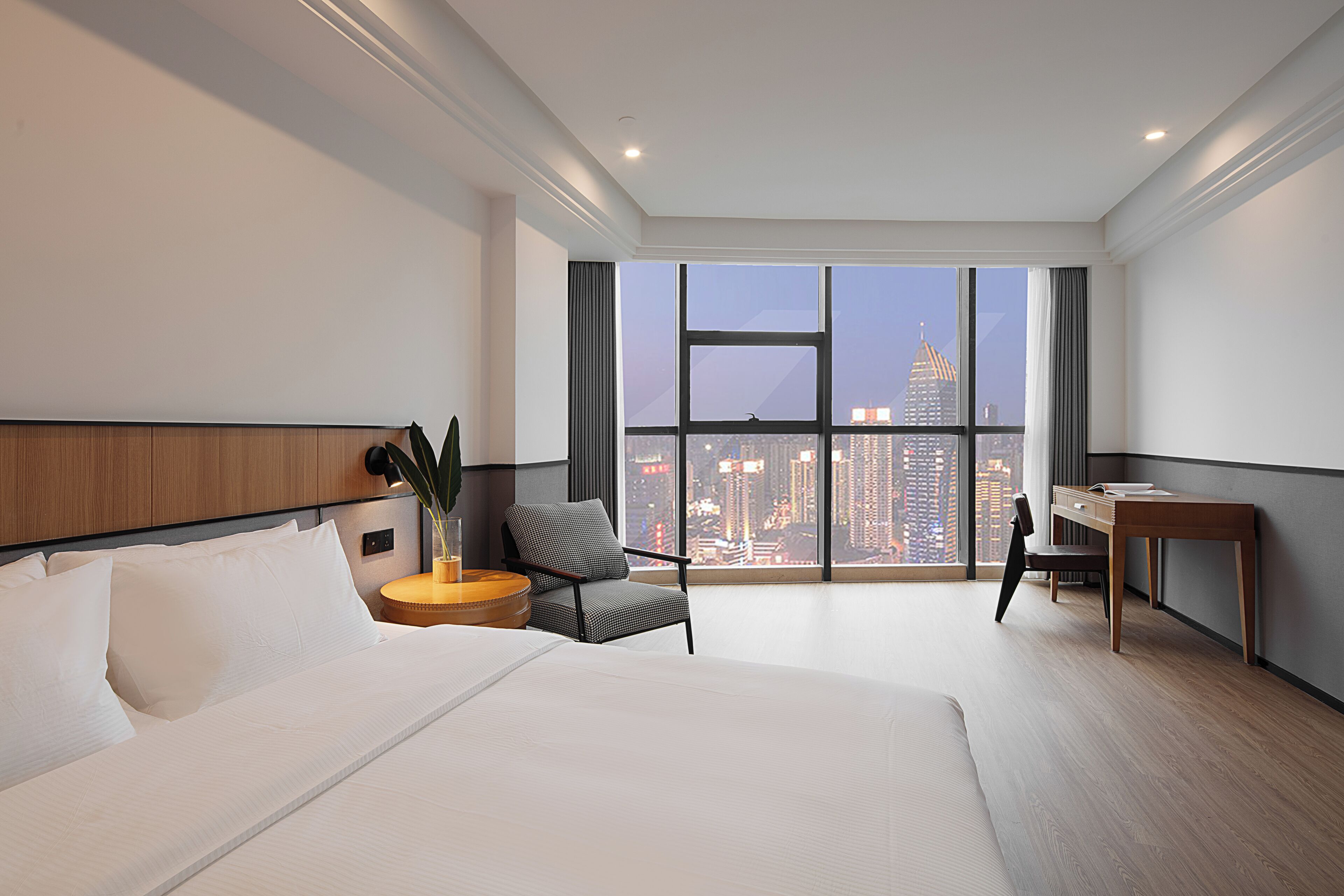 A modern hotel room with a large bed, a desk, a chair, and a view of the cityscape through large windows.