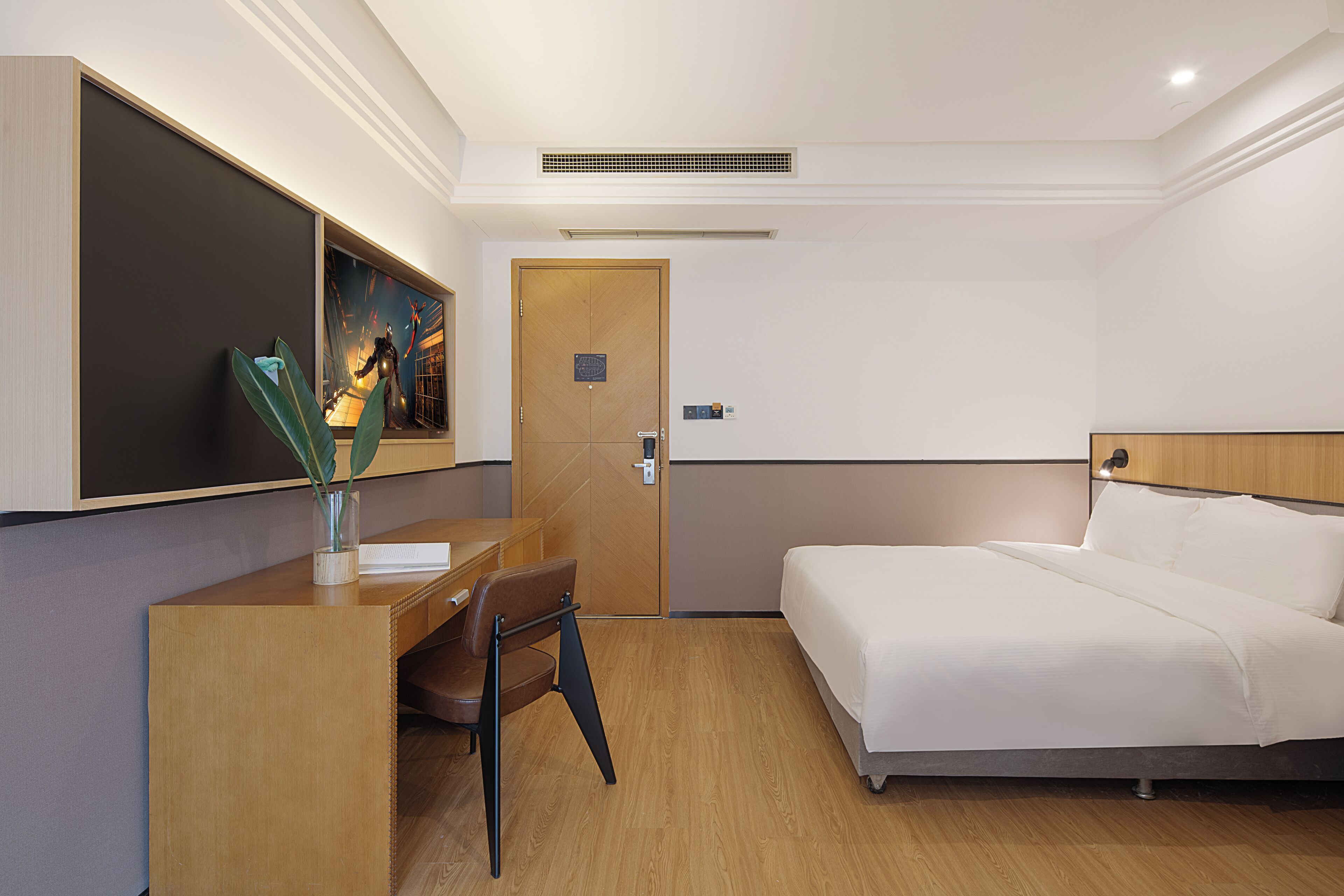 A hotel room with a bed, a desk, and a chair. There is a painting on the wall, a door, and a vent on the ceiling.