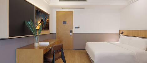 A hotel room with a bed, a desk, and a chair. There is a painting on the wall, a door, and a vent on the ceiling.