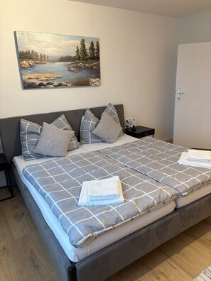 A double bed with a checkered bedspread, two pillows, and a folded towel. There is a nightstand with a lamp and a painting of a landscape on the wall.