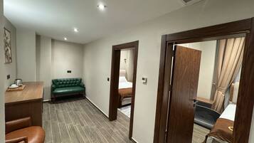 Habitación de hotel con escritorio de madera, sofá verde, sillón de cuero marrón y cama con manta marrón.