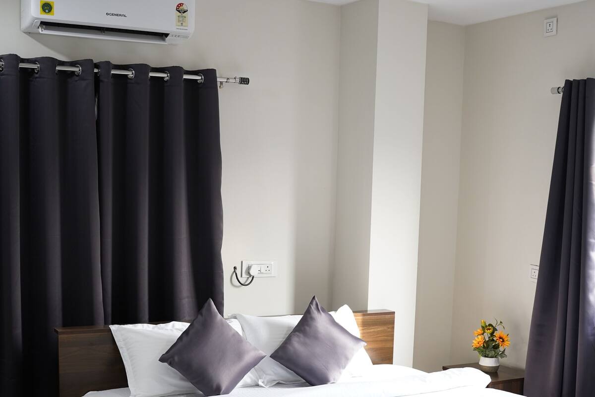 Ein Hotelzimmer mit einem Bett, dunklen Vorhängen, einer Klimaanlage, einem Nachttisch mit Blumen und einem Deckenventilator.
