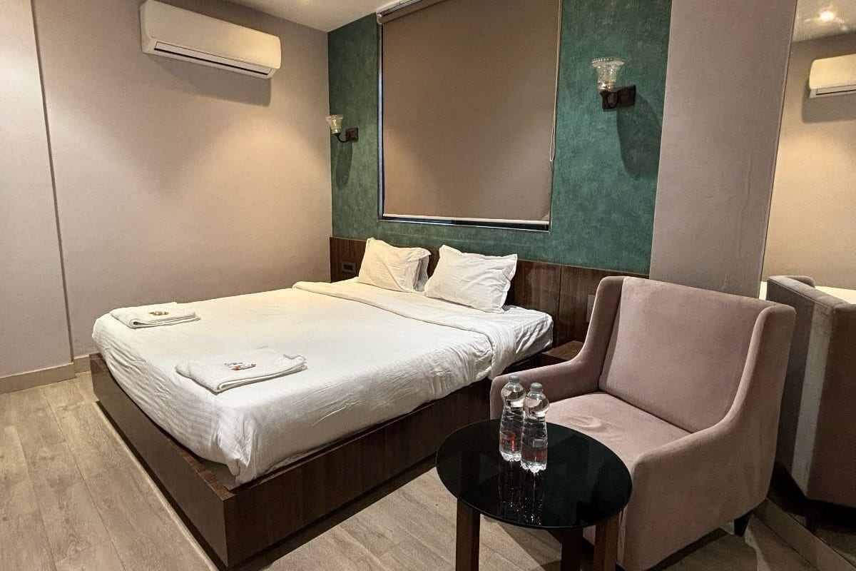 A modern hotel room with a bed, a chair, a small table with water bottles, a ceiling fan, and an air conditioning unit.