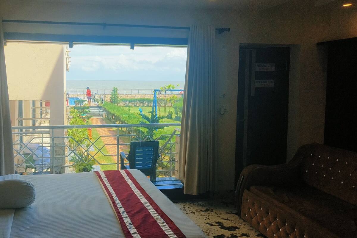 A hotel room with a large bed, a sofa, and a balcony with a view of the beach and palm trees.