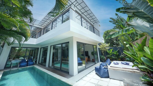 Una casa moderna de dos pisos con balcón de vidrio, piscina y exuberante vegetación tropical.