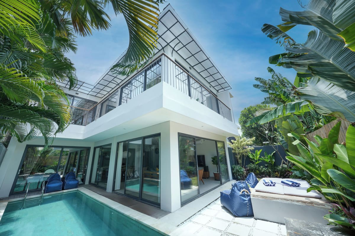 A modern two-story house with a glass-enclosed balcony, a swimming pool, and lush tropical vegetation.