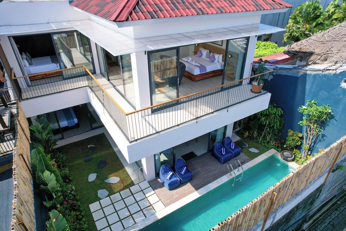 A modern two-story house with a balcony, a pool, and lush greenery.