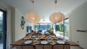 Comedor con una gran mesa dispuesta para una comida, rodeada de sillas de mimbre, y vistas a una piscina y vegetación al exterior.