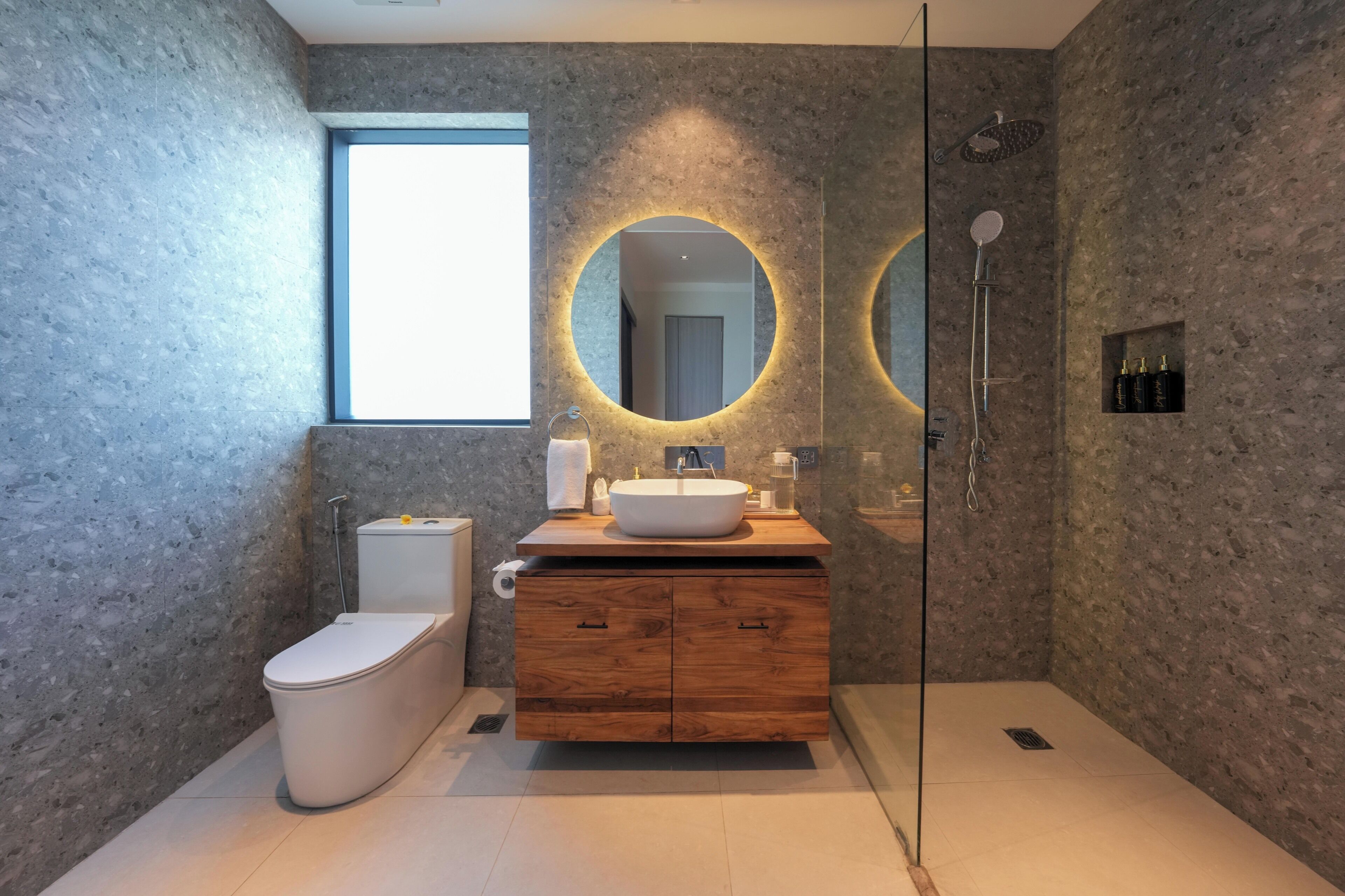 Une salle de bain moderne avec un mur en pierre, un grand miroir rond avec éclairage intégré, un meuble-lavabo en bois, des toilettes et une douche.