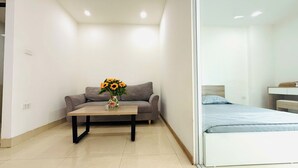 A modern room with a grey sofa, a wooden coffee table with a vase of flowers, a bed with a blue mattress, and a white bedside table. - Era Apartment Dinh Thon (Hanoi)