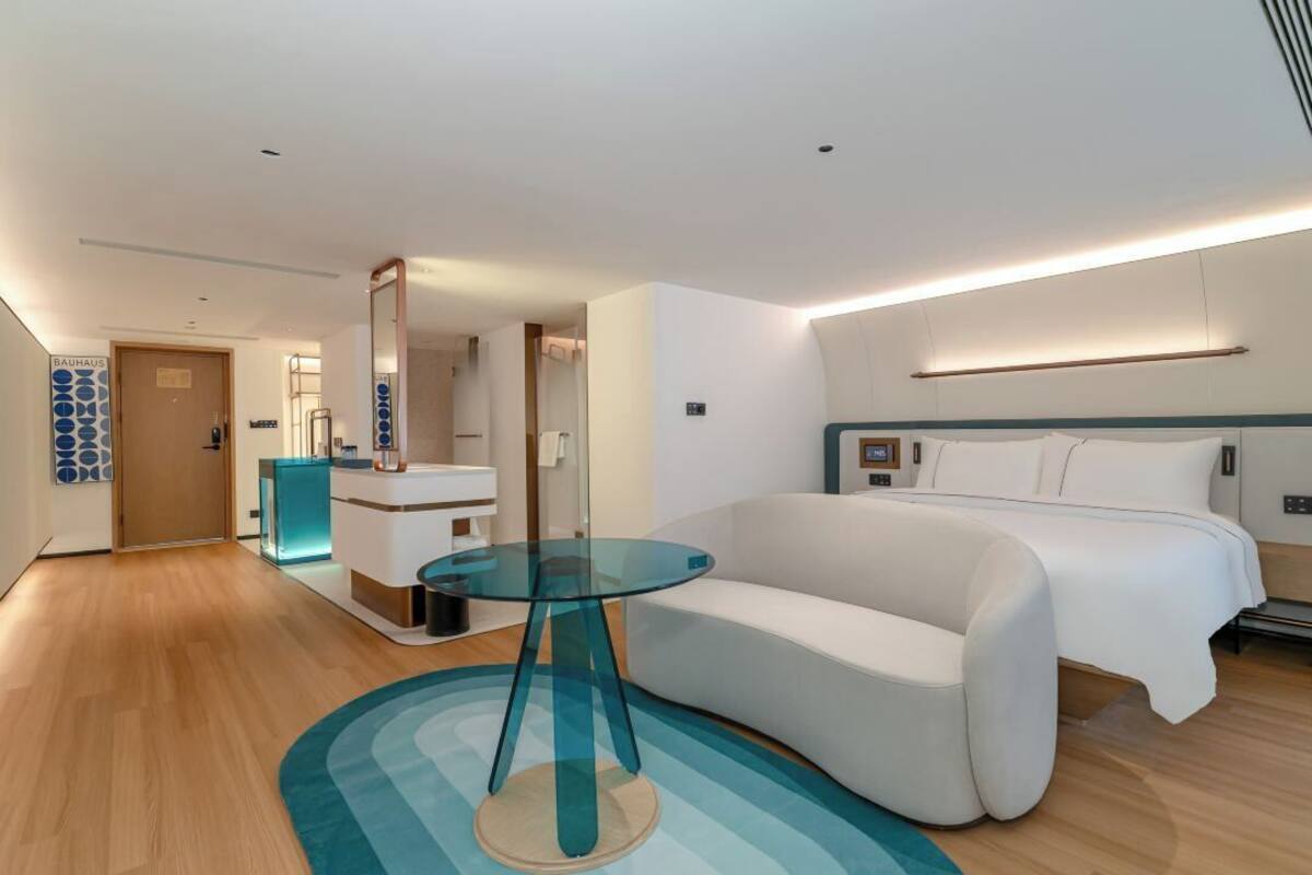 Una habitación de hotel moderna con una cama grande, una mesa de vidrio, un sofá blanco y un baño con un detalle en tono azul.