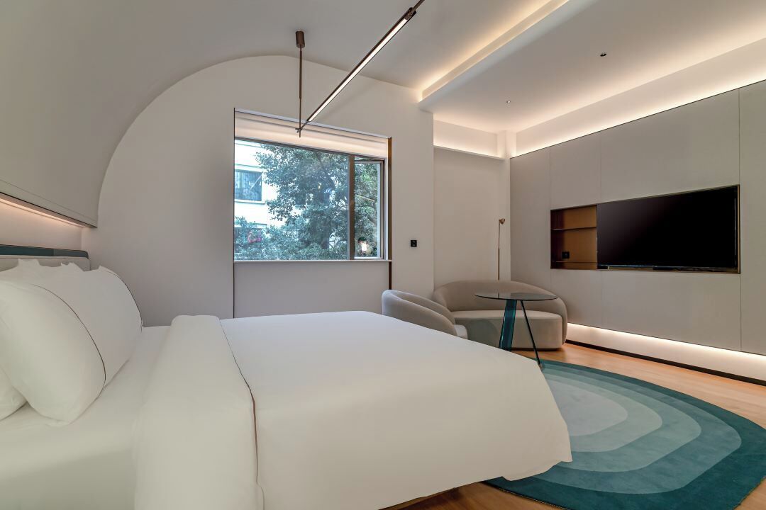 Un dormitorio moderno con una cama grande, un televisor en la pared, un techo curvo y vistas a árboles desde el exterior.