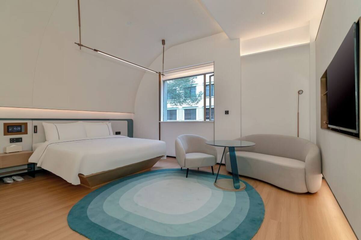 Una habitación de hotel moderna con una cama, un sofá, una silla, una mesita pequeña y un ventanal grande.