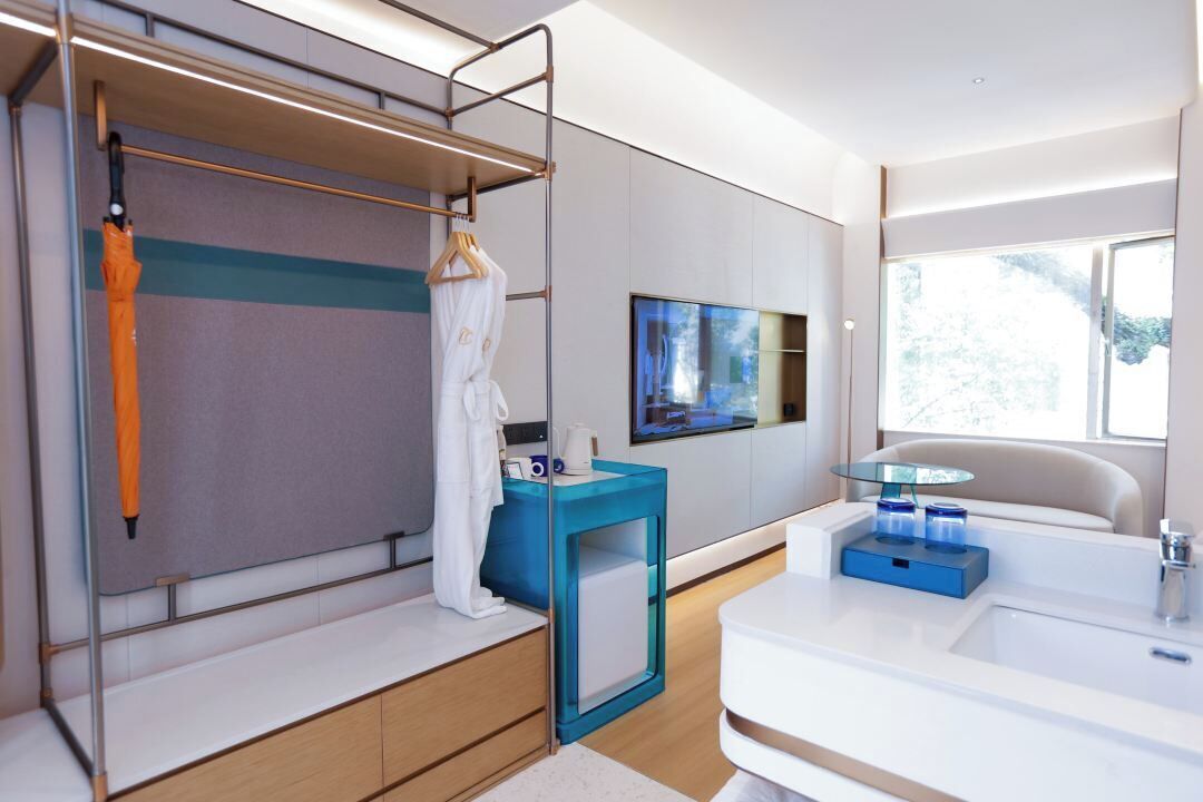 A modern hotel room with a bunk bed, a bedside table with a glass top, a flat-screen TV mounted on the wall, and a large window providing natural light.