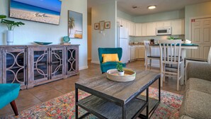 A living room with a TV displaying a beach scene, a teal armchair, a wooden coffee table, and a kitchen area with white cabinets and a microwave. - Port A Golf Cart Zone • Shared Pool + Pet-Friendly | Costa Bahia (Port Aransas)