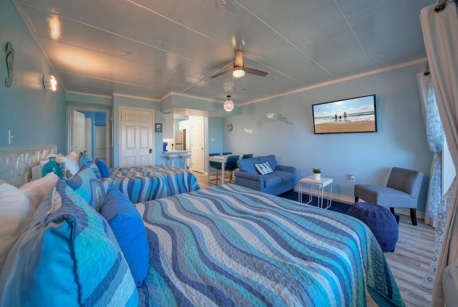 A hotel room with two beds, a desk, a chair, a small table, a ceiling fan, and a beach scene on the wall.