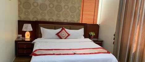 A neatly made bed with white bedding and a red and white decorative runner. There is a nightstand with a lamp and a small potted plant beside the bed. The room has a patterned wallpaper and a window with blinds.