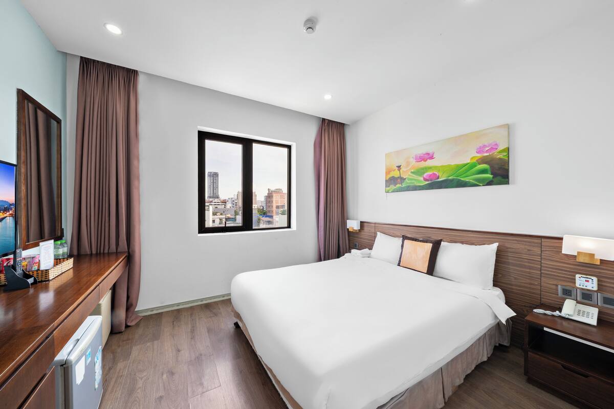 A hotel room with a large bed, a desk with a computer, a telephone, and a window with a city view.