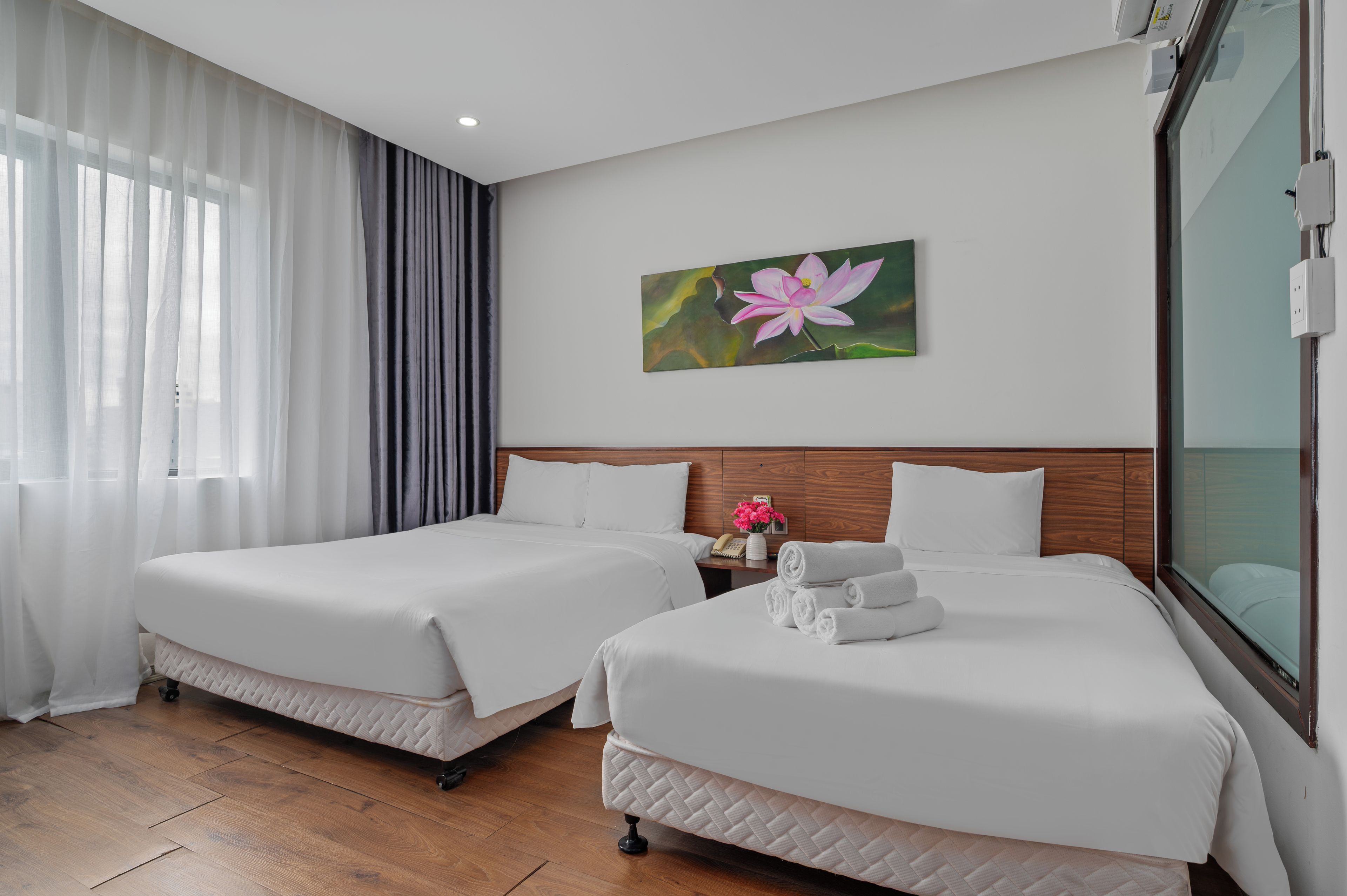 A hotel room with two beds, a painting of a flower on the wall, and a window with curtains.