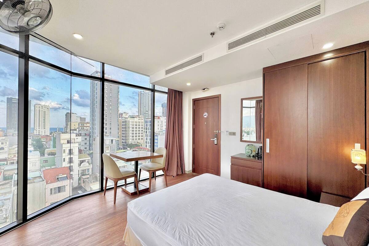 A modern hotel room with a large window offering a city view, a bed, a desk with a chair, and a wardrobe.