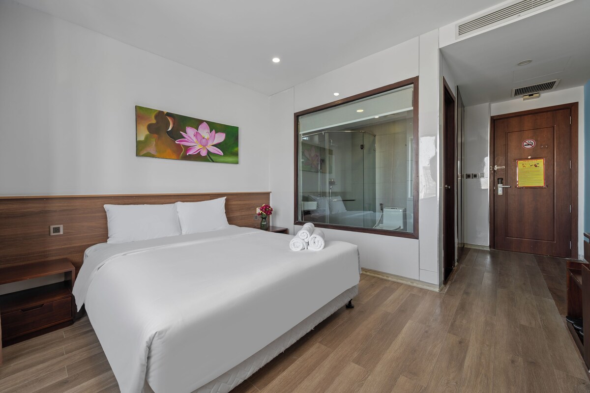 Habitación de hotel con una cama grande, un cuadro de una flor, un box de ducha de vidrio y una puerta de madera.