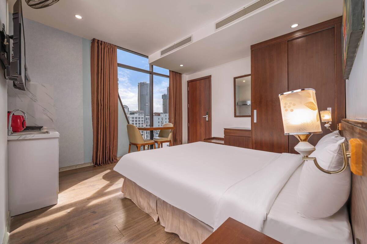 A hotel room with a bed, a desk with a red kettle, a chair, a window with a city view, and a lamp.
