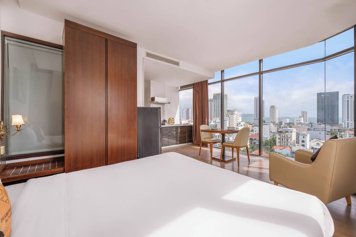 A hotel room with a large bed, a desk with chairs, a view of the city through large windows, and a sliding door.