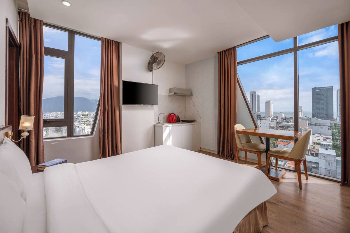 A hotel room with a bed, a desk, a chair, a TV, and a view of the city.