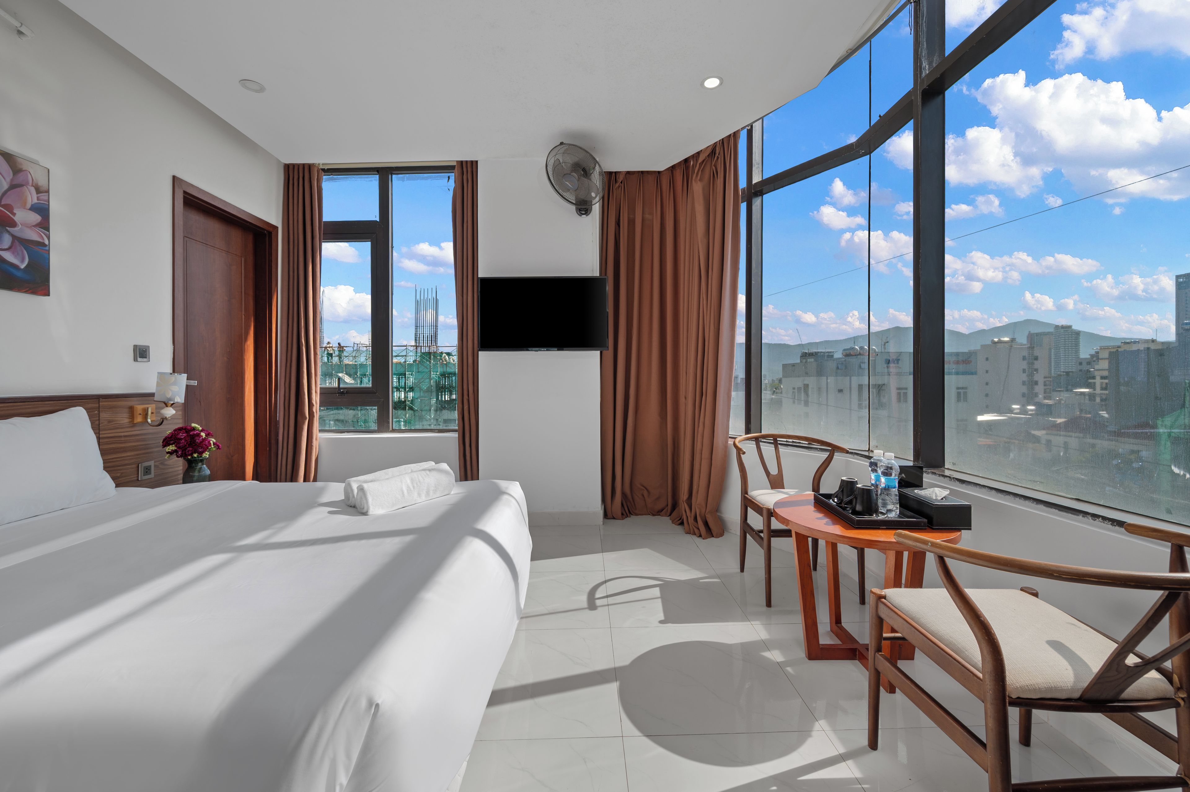 Quarto de hotel com uma cama grande, uma mesa com cadeira, uma janela com cortinas e vista para a paisagem urbana.