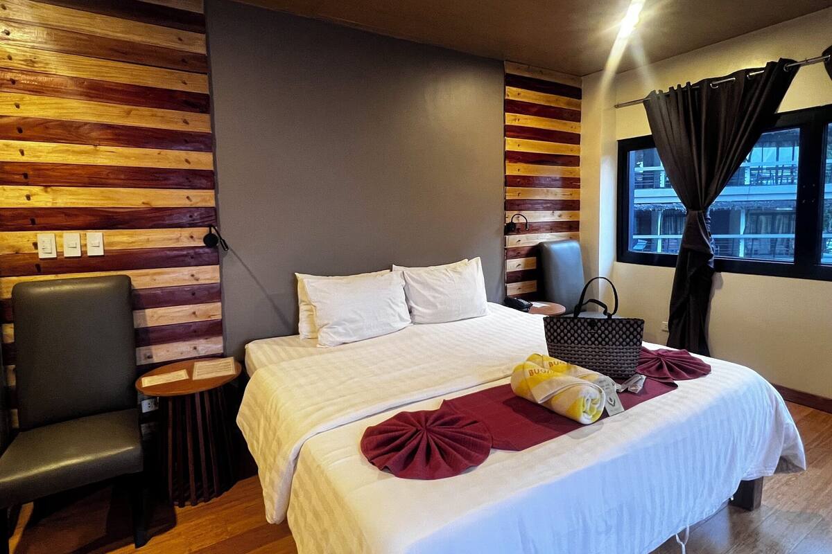 Habitación de hotel con cama, una silla, mesita de noche, ventana con cortinas y un panel de pared de madera.