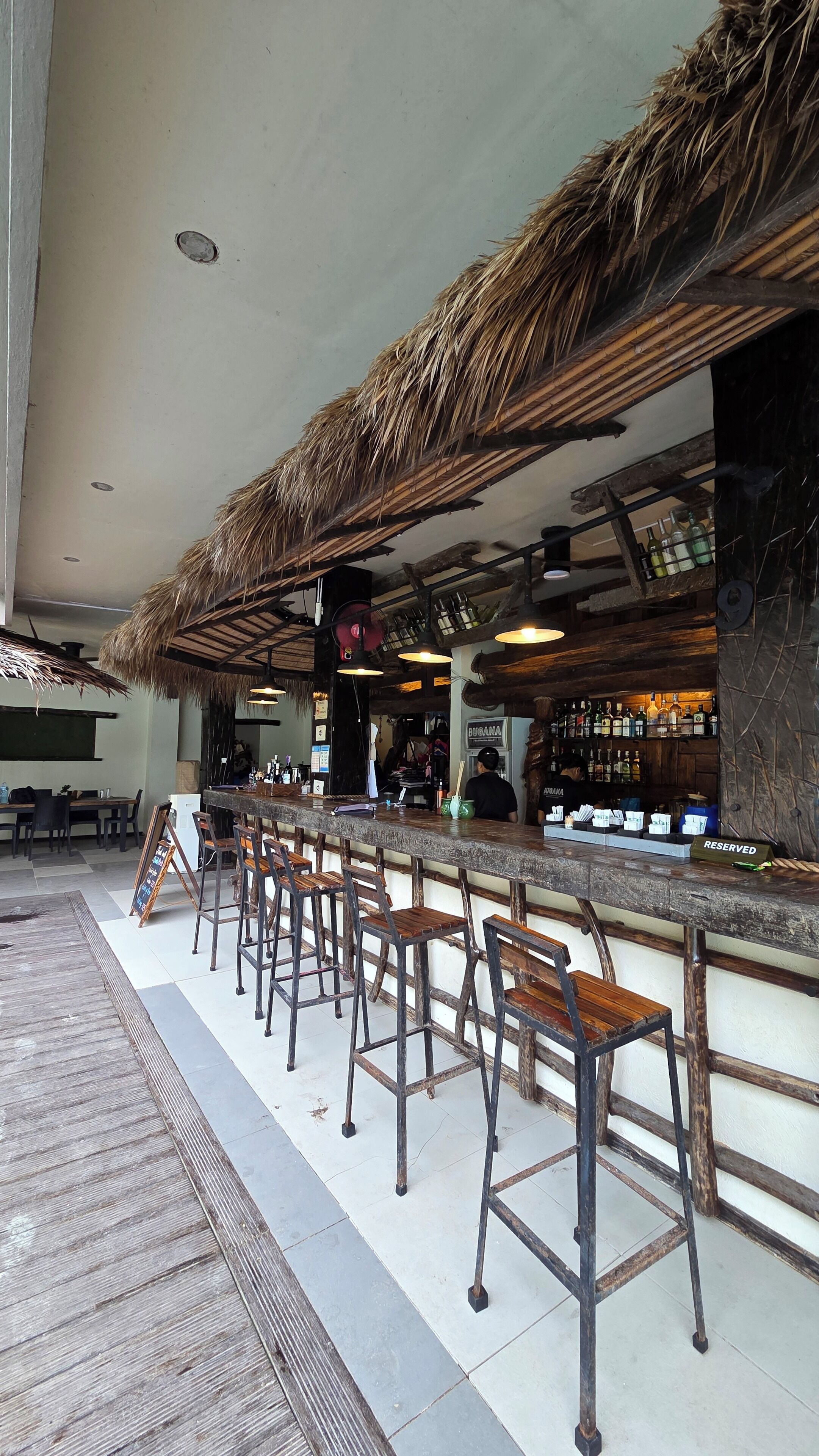 Zona de bar con techo de paja, taburetes de madera y mostrador con botellas.