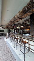 Zona de bar con techo de paja, taburetes de madera y mostrador con botellas.