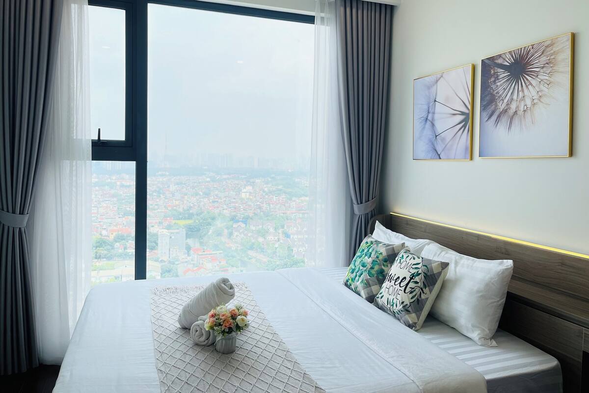 Habitación de hotel con un amplio ventanal que ofrece vistas a la ciudad, una cama bien hecha con toalla y flores, y dos cuadros en la pared.
