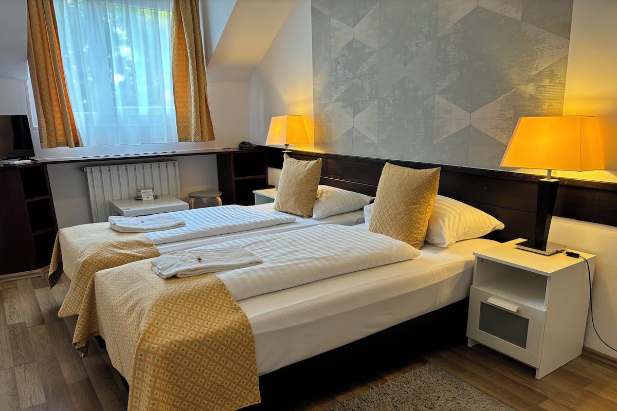 A hotel room with two beds, a bedside table with a lamp, a small desk, and a radiator.