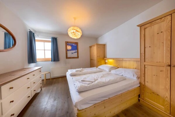 A bedroom with a wooden bed, a wooden wardrobe, a bedside table, a mirror, and a window with curtains.
