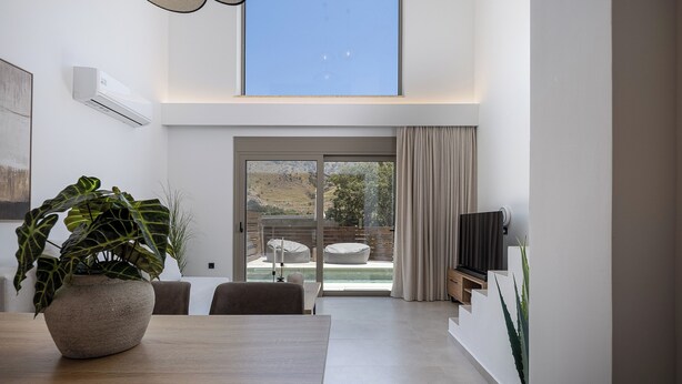 A modern living room with a large window, a dining area with a table and chairs, and a TV mounted on the wall. - Imbros Villas Free* Heated Pools in mountains! (Crete)