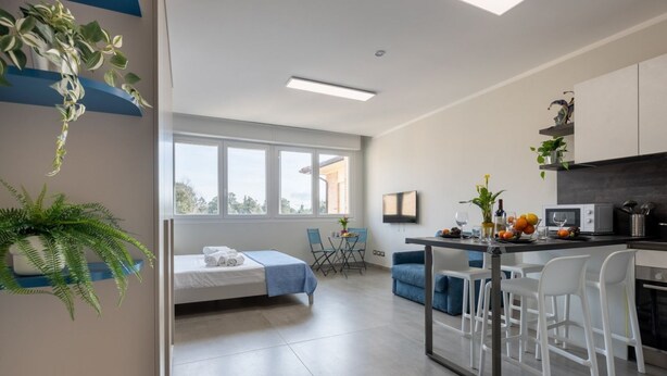 A modern hotel room with a bed, a dining area, a kitchen, and a large window.