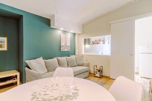A modern living room with a teal accent wall, a grey sofa, a white dining table, and a small side table with a lamp.