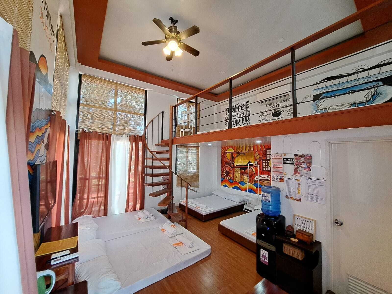 A compact hotel room with a bunk bed, a ceiling fan, a window with curtains, a wall with a coffee advertisement, and a welcome sign.